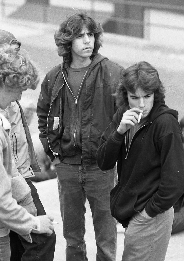 Black-and-white photo of young men standing outdoors. The man in the center wears a short zip-front jacket over a hooded sweatshirt