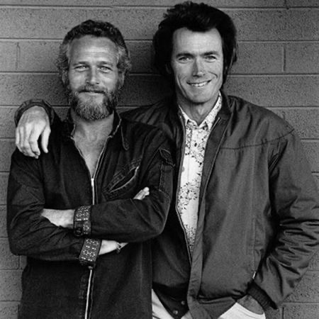 Black-and-white photo of two men in front of a brick wall. The man on the right wears a short jacket over a patterned shirt