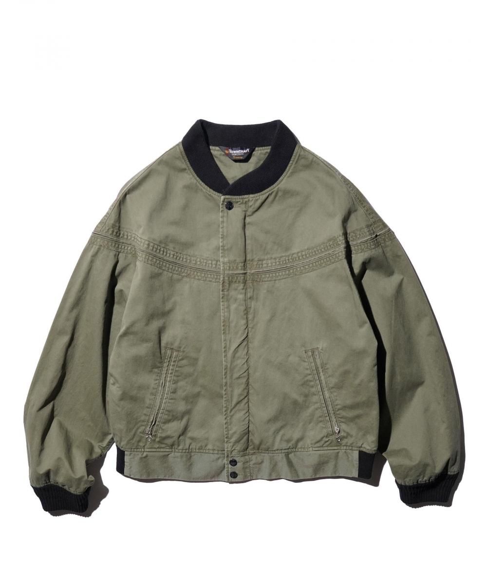 Front of the pigment-dyed derby jacket in OLIVE. Black rib collar and cuffs, chest seam line with decorative stitching, slanted zip pockets on the left and right at the waist